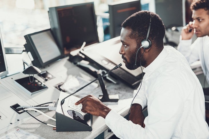 A man in a control room watches a monitor and speaks into a microphone